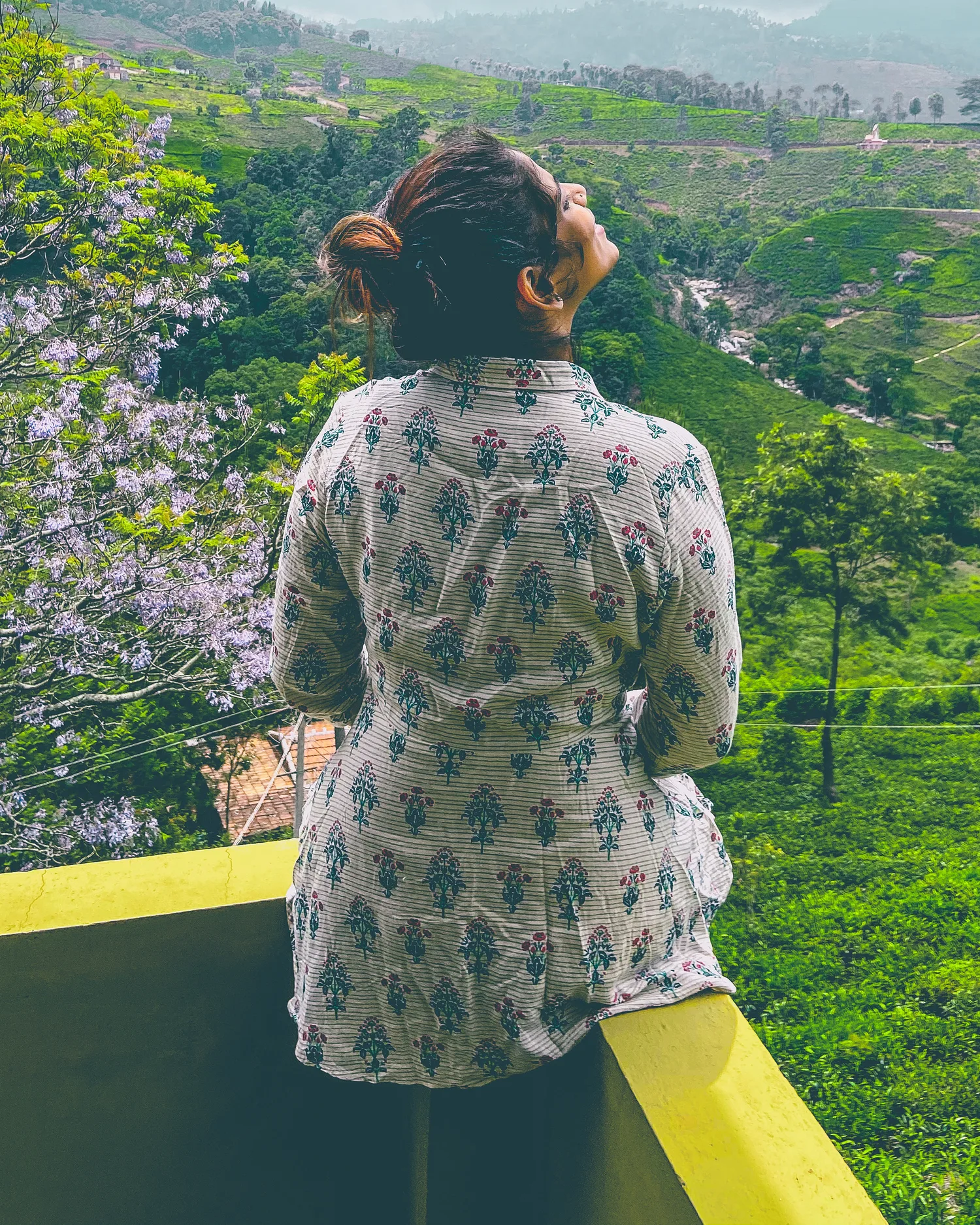 A woman seated on a ledge with her back to us, looking out over misty tea hills.